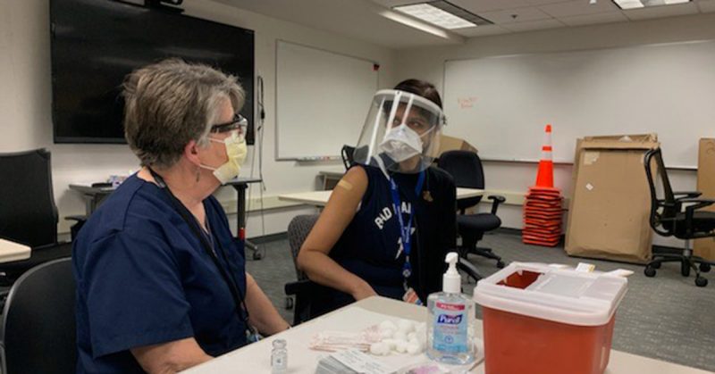 two people sitting at a table in a media setting- one with a mask on and the other has a full face shield