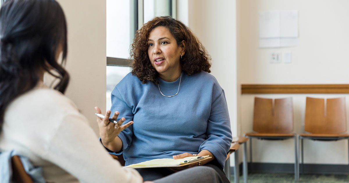 woman in a blue sweater holding a clipboard speaking to person in white sweater with her back to camera