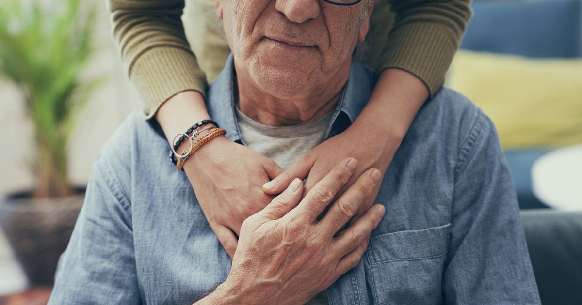 woman stands behind seated older man with her arms clasped around his front