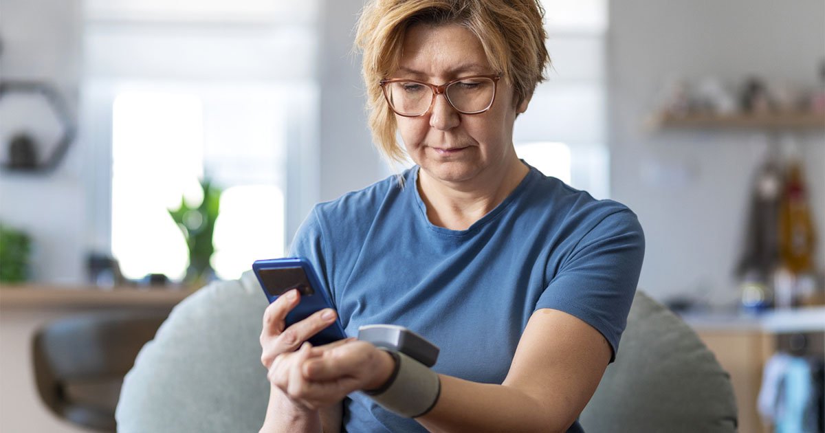 Older woman sitting at home using a cell phone and blood pressure device attached to her wrist to monitor her blood pressure