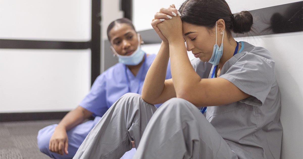 A distressed female healthcare worker is sitting in a hospital hallway with her head in her hands. A female colleague sits next to her.