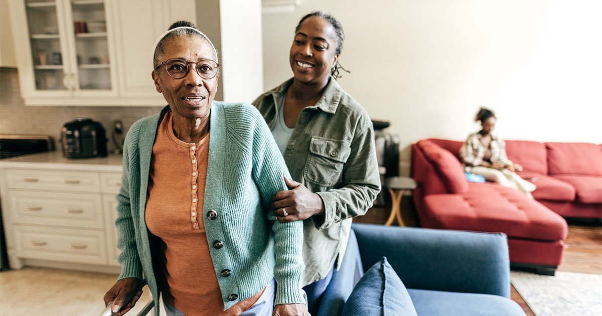 Older Black woman being helped by middle aged Black woman to walk in a living room