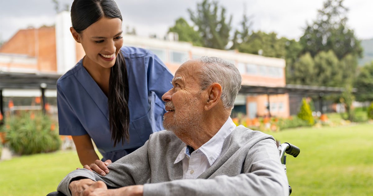 Female care provider in a blue uniform speaks to an older man seated in a wheelchair